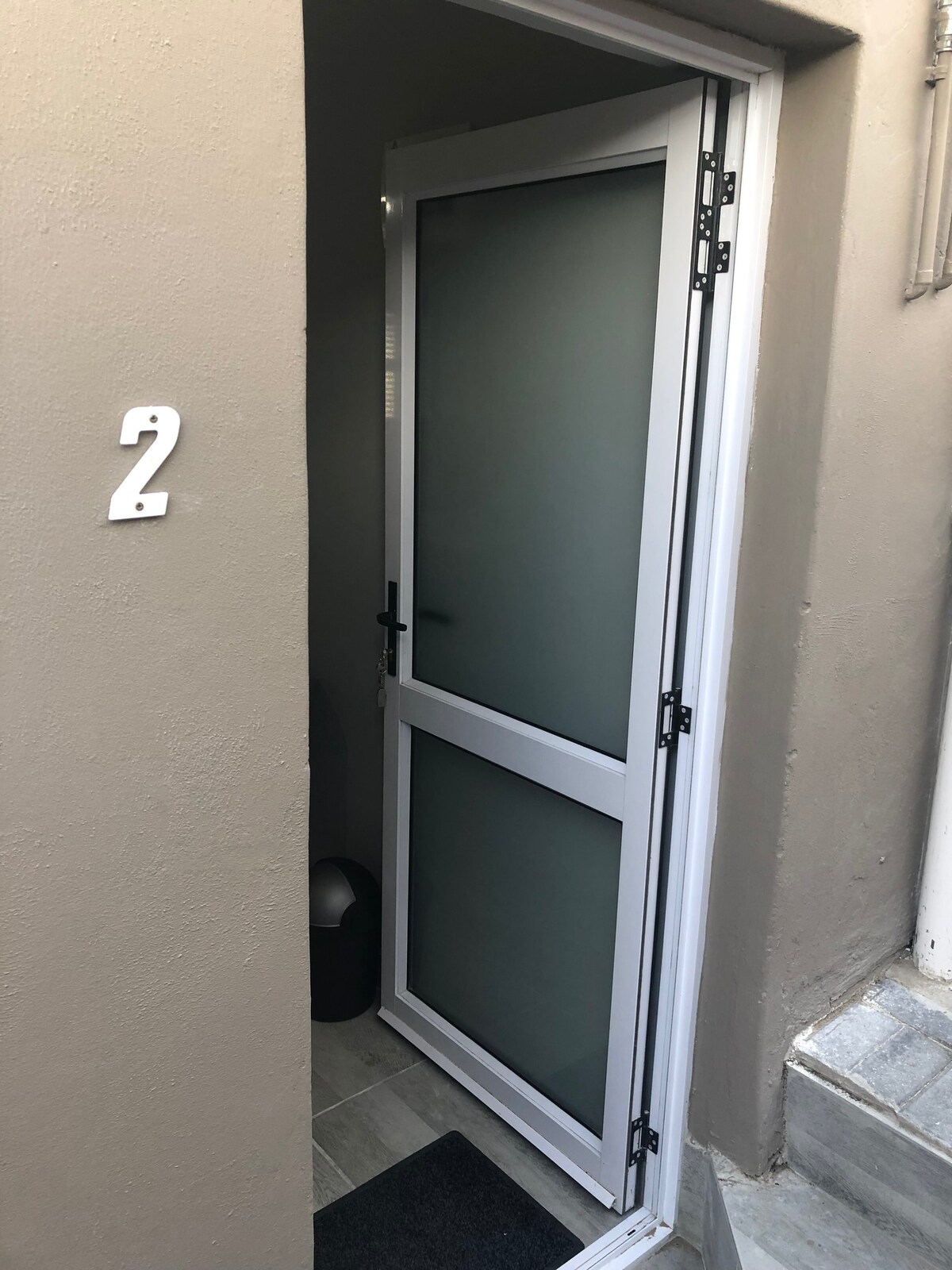 A modern entrance door is visible, featuring a white frame and frosted glass panels. The door opens to a small entryway, with a doormat positioned on light-colored flooring. A number '2' is displayed on the wall beside the door.