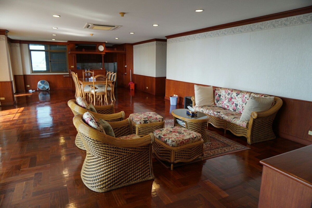 A spacious living area features wooden flooring and light-colored walls. Comfortable seating includes a sofa and armchairs adorned with floral patterns. A wooden dining table is visible in the background, while a clock is mounted on the wall. Large windows allow natural light into the room.