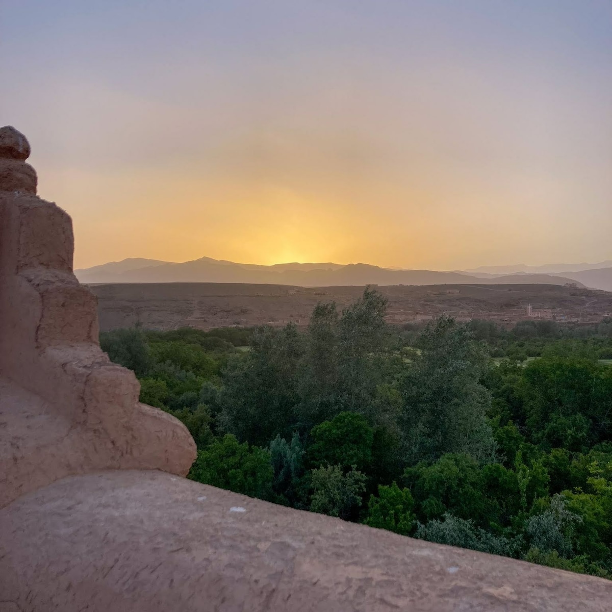 The image captures a serene sunset view over the valley, with soft orange and yellow hues blending into the sky. Lush green trees and distant mountains provide a tranquil backdrop, framed by the textured wall of the terrace, inviting a sense of calm.