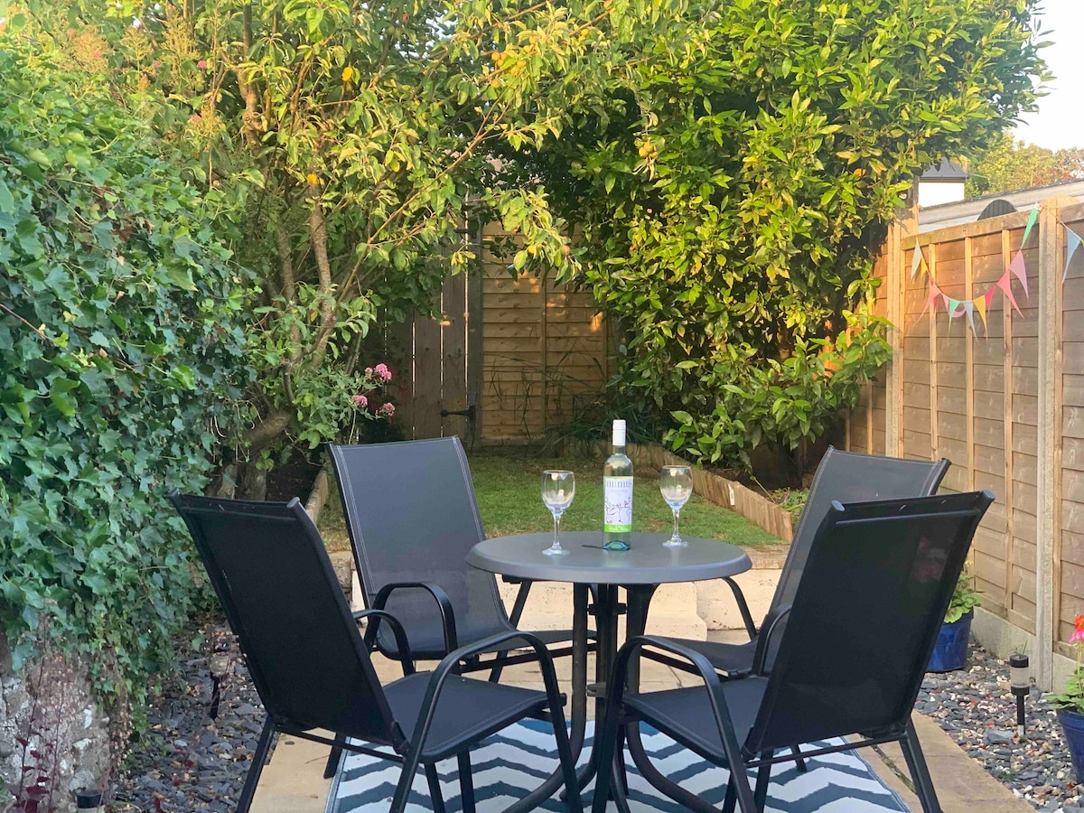 A private enclosed garden is showcased, featuring a circular table surrounded by four black chairs. A bottle of wine and two glasses sit atop the table. Lush greenery and established shrubs provide a serene backdrop, while a narrow lawn area can be seen beyond the patio.