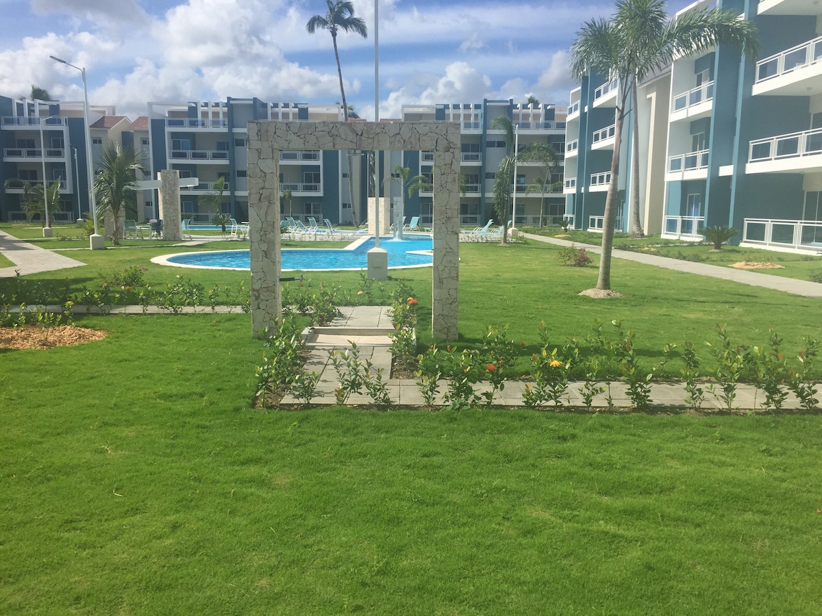 An outdoor view of the condo complex is presented, featuring well-maintained green lawns and neatly arranged flower beds. In the background, a swimming pool is visible, surrounded by lounge chairs and palm trees, contributing to a refreshing and inviting environment.