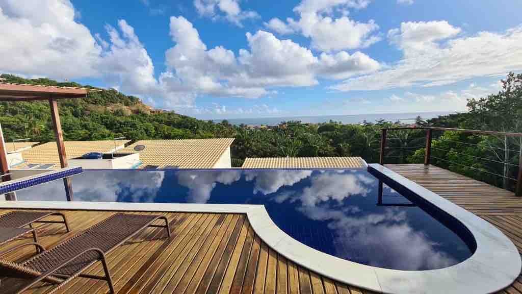 An infinity pool is seen with a wooden deck, surrounded by lush greenery. The shimmering water reflects the sky, featuring white clouds and a panoramic view of the landscape beyond.