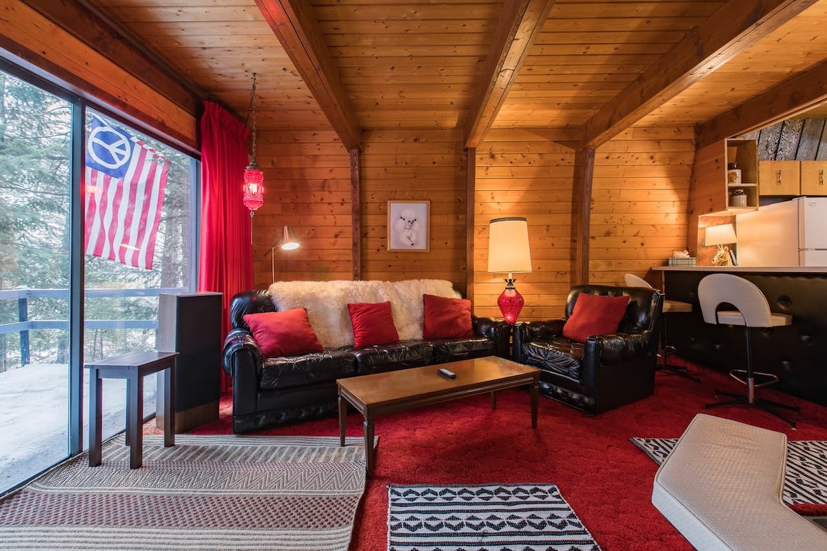 A cozy living area features a plush black leather couch adorned with bright red pillows and a soft white throw. Natural wood paneling and exposed beams create a warm ambiance. A vibrant red rug complements the space, which includes a coffee table and a large window with an outdoor view.