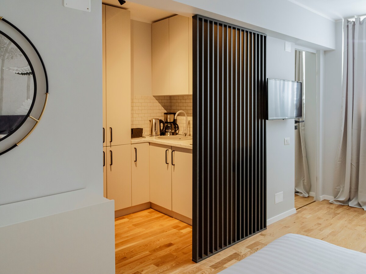 A kitchenette is visible, featuring sleek cabinetry and a variety of appliances, including a coffee maker. A television is mounted on the wall across from the sleeping area. Natural light enters through sheer curtains, illuminating the warm wooden flooring throughout the space.