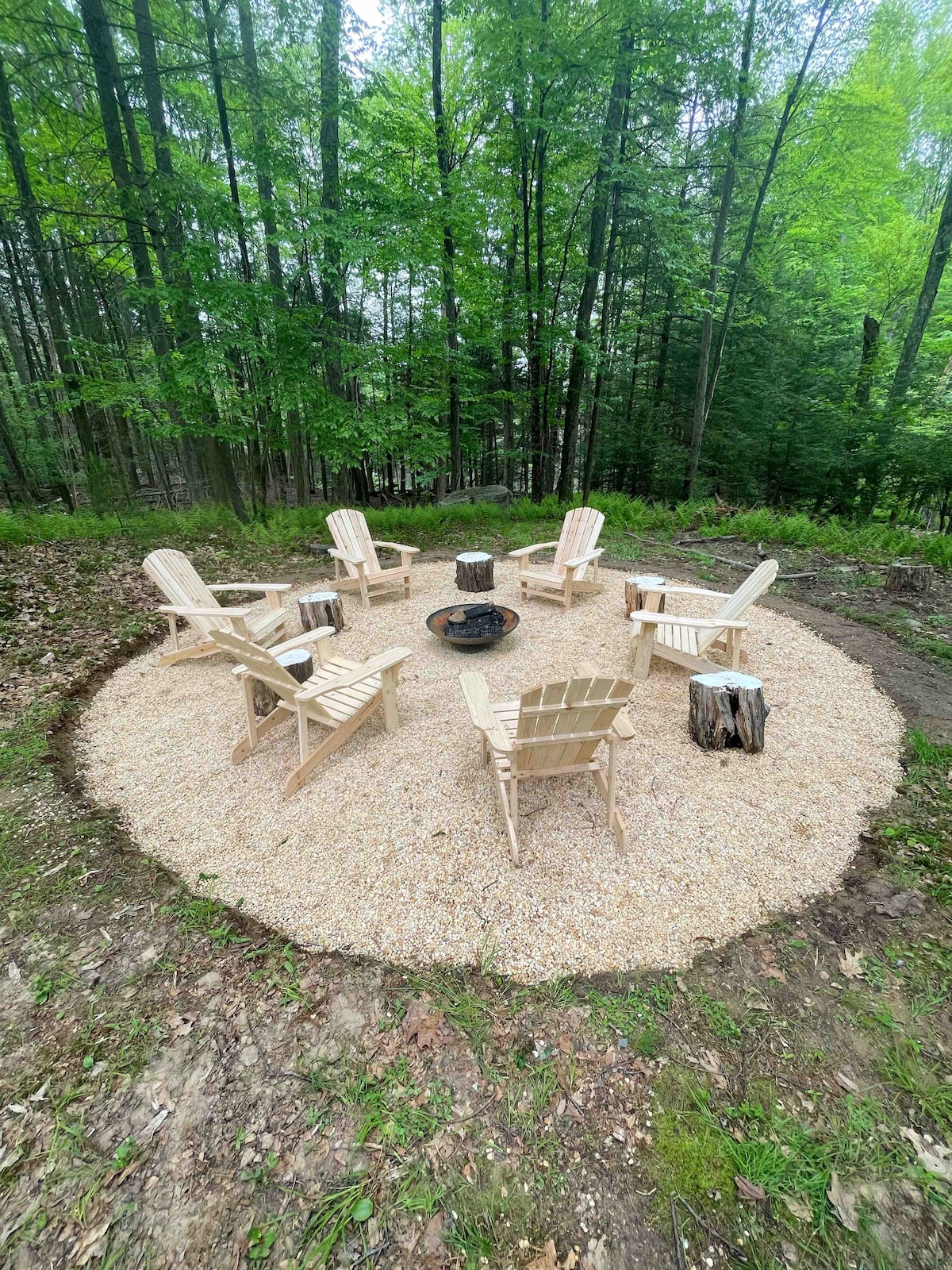 A fire pit area is surrounded by six light-colored Adirondack chairs arranged in a circular formation. The ground is covered with small gravel stones, and nearby natural greenery creates a serene setting in the woods.