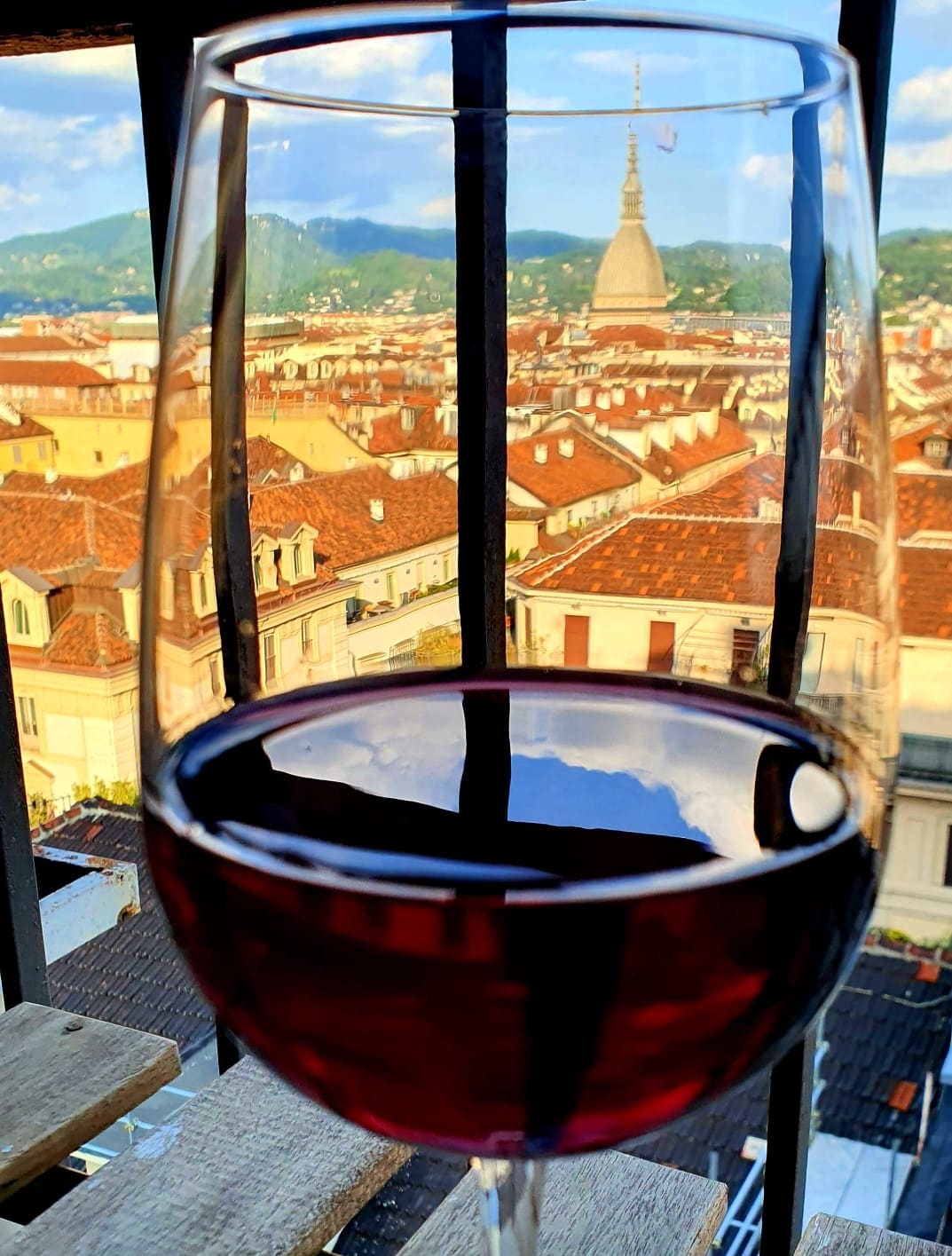 A larger wine glass filled with deep red wine is positioned in front of a panoramic view of Torino. The glass reflects the surrounding rooftops and the iconic Mole Antonelliana, providing a glimpse of the city's terracotta roofs and vibrant skyline.