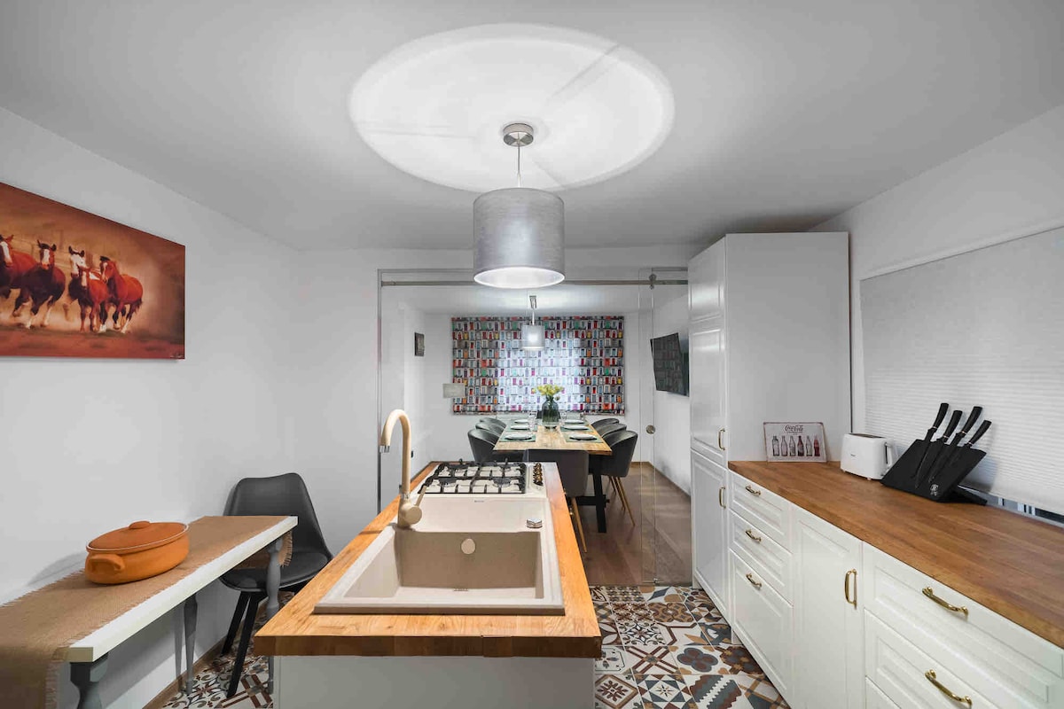 A modern kitchen is highlighted by a central island featuring a gas cooktop. Cabinets in a light finish offer ample storage. A dining table is positioned nearby, adorned with colorful decor, while a window dressed in patterned curtains allows natural light to filter in.
