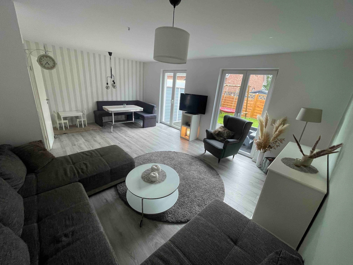 A spacious living area features comfortable seating arranged around a circular coffee table. A large window allows natural light to fill the space, while a cozy rug adds texture. A flat-screen TV is mounted on the wall, and a dining table is visible in the background.