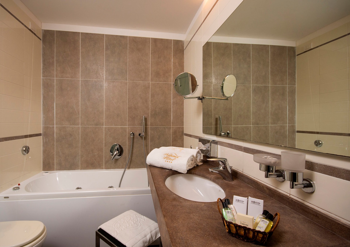 The bathroom features a modern design with a spacious vanity area and a large mirror. A private jacuzzi tub is situated beside the shower, while toiletries and amenities are neatly arranged on the counter, adding convenience for guests.