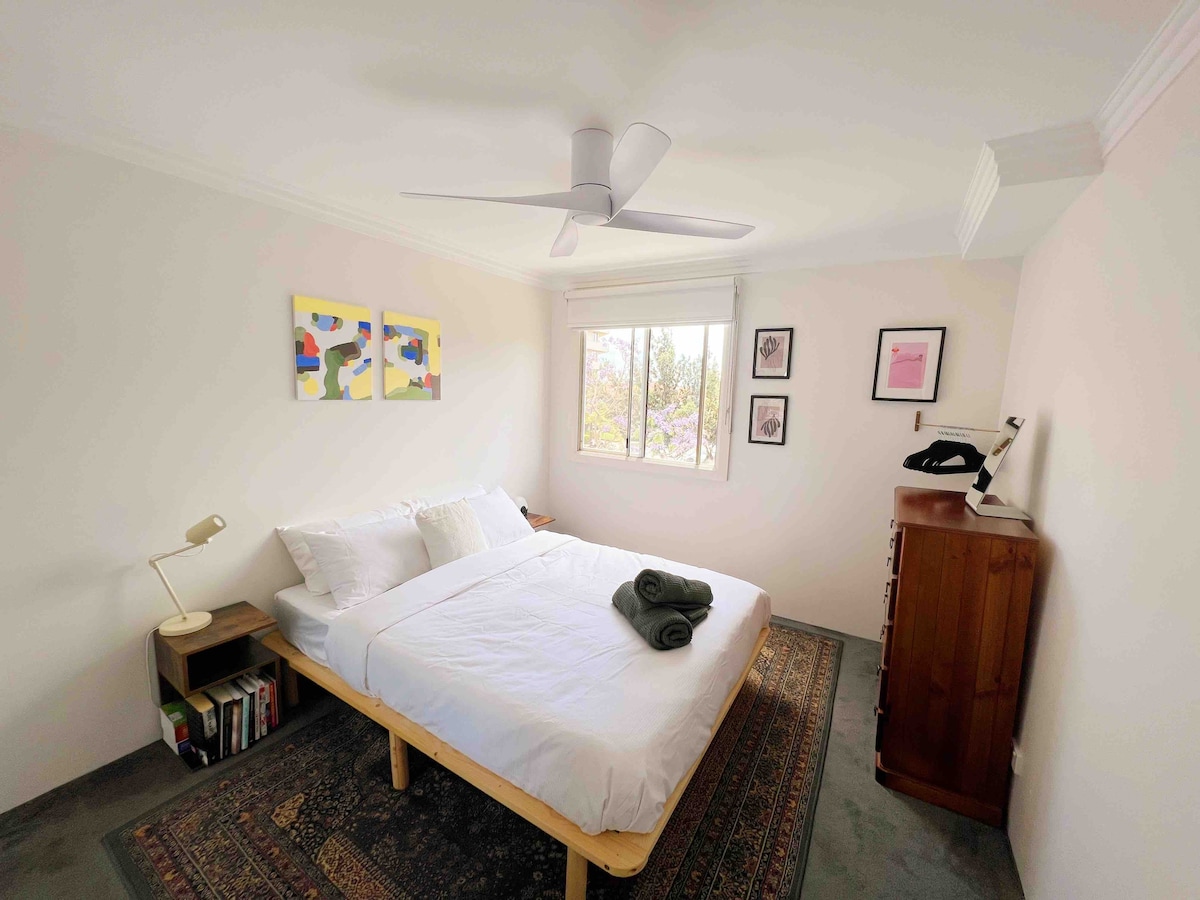 A well-lit bedroom features a spacious bed adorned with fresh linens and neatly rolled towels. A ceiling fan offers air circulation, while artwork is displayed above the bed. Natural light fills the room through a window, illuminating a wooden dresser and bedside table.