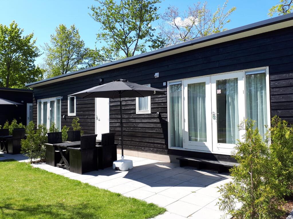 A modern chalet exterior is observed, featuring dark wooden paneling and large windows with white drapes. An outdoor seating area with a table and chairs, accompanied by a black umbrella, offers shade. Lush greenery and well-maintained grass enhance the inviting atmosphere.