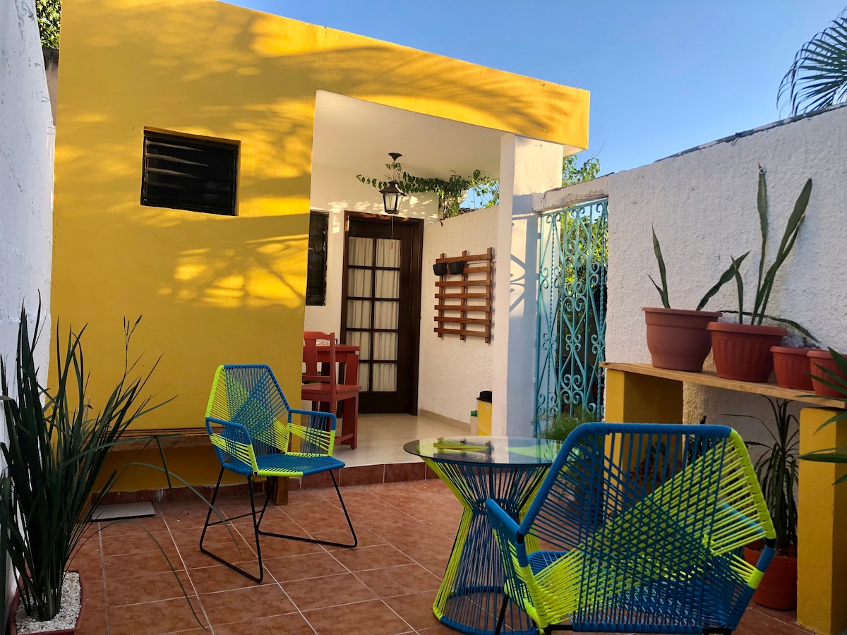 A vibrant outdoor terrace showcases a bright yellow wall and terracotta tiles. Two stylish, colorful chairs and a glass table are surrounded by plants in pots, creating a refreshing space for relaxation.