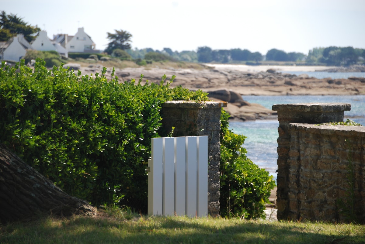 A white wooden gate stands between two stone pillars, leading to a serene coastal view. Lush green foliage frames the entrance, with rocky shores and distant houses visible by the water under clear skies.