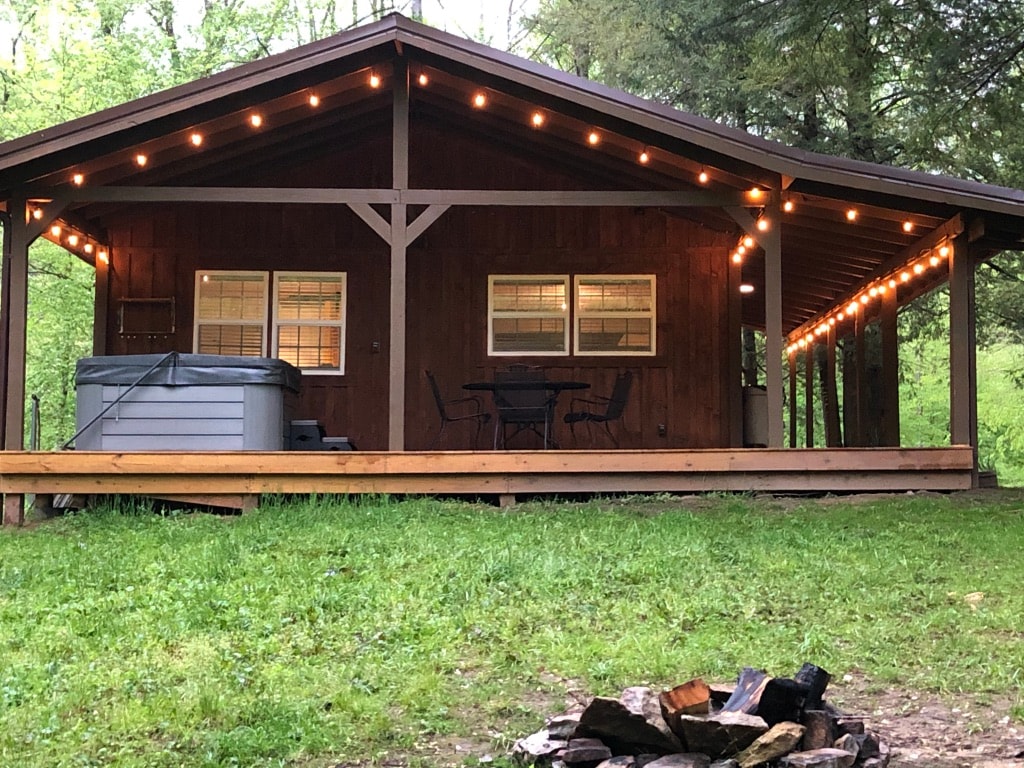 A rustic cabin is set amid lush greenery, featuring a spacious deck with outdoor lighting. A hot tub is positioned nearby, while a small table and chairs are arranged for seating. A fire pit with logs is visible in the foreground, enhancing the outdoor charm.