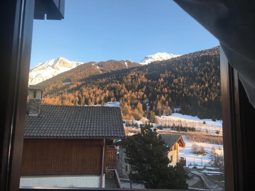 A scenic view is captured from a window, showcasing a mountainous landscape adorned with trees. Snow covers lower areas, while the peaks are capped with snow. Nearby rooftops offer a glimpse of the alpine architecture, with clear blue skies enhancing the natural beauty.