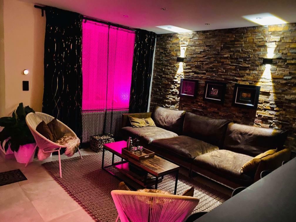 A stylish living area features a brown sofa positioned against a textured stone wall. Soft lighting creates a cozy ambiance, while a bright pink curtain adds a pop of color. A rectangular coffee table sits in front of the sofa, surrounded by tasteful decorations.