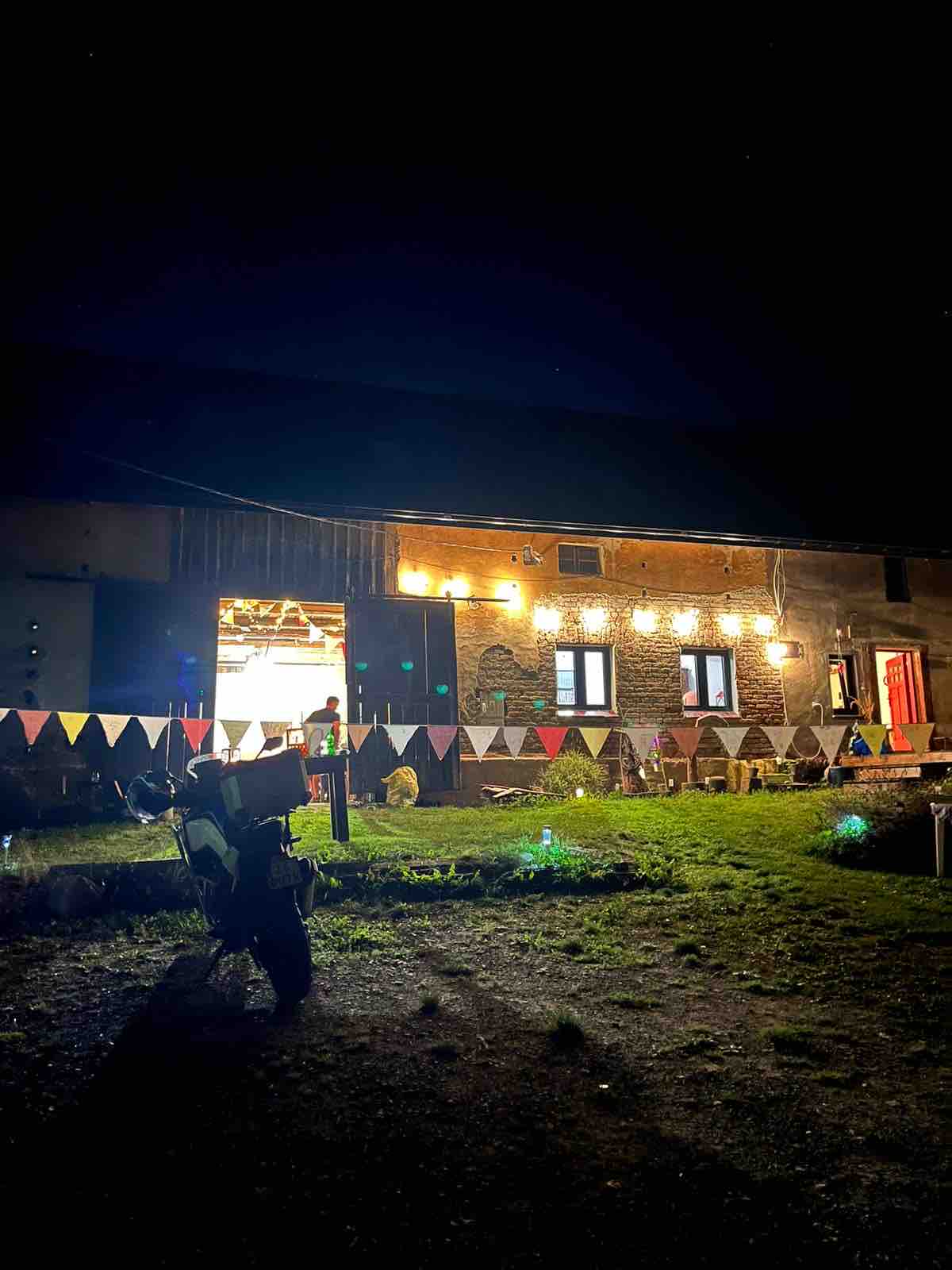 The exterior of a cozy farmhouse is illuminated at night, featuring string lights that enhance its welcoming ambiance. Colorful bunting decorations are displayed along the facade, adding a festive touch. A person is seen standing in front of the entrance, surrounded by a lush grassy area.