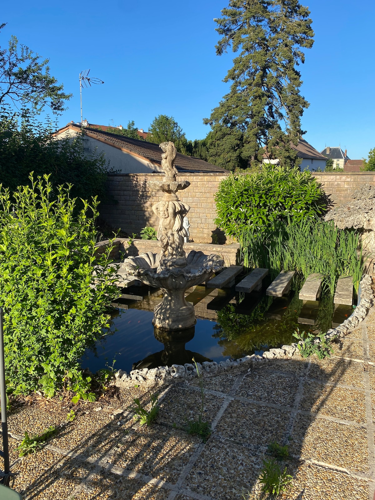 A peaceful garden scene features a central ornamental fountain surrounded by water. Lush greenery and tall plants provide a natural frame, while a stone pathway leads to the fountain, suggesting a space for relaxation. Sunlight illuminates the tranquil setting.