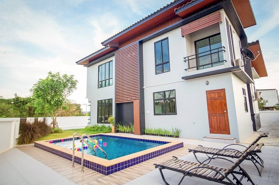 A modern house features large windows and a wooden door, showcasing a private swimming pool adorned with colorful pool floats. Wooden loungers are positioned beside the pool, creating an inviting space for relaxation. Lush greenery surrounds the property, enhancing the tranquil outdoor setting.