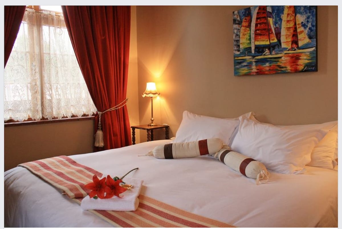 A spacious bed is adorned with neutral linens and decorative pillows, complemented by a vibrant artwork featuring sailboats. A small side table with a lamp provides gentle illumination, while sheer curtains frame the window, allowing soft light to enter the room.