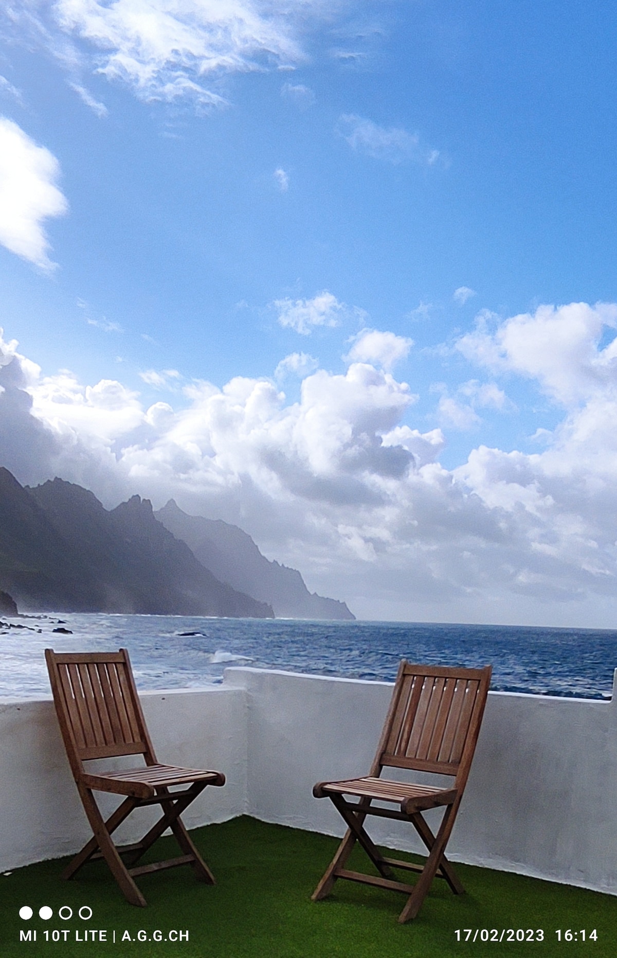 A serene terrace is shown with two wooden chairs positioned to face the Atlantic Ocean. Rolling hills and dramatic cliffs frame the background, while a partly cloudy sky adds depth to the view. The sound of waves can be imagined in this tranquil setting.