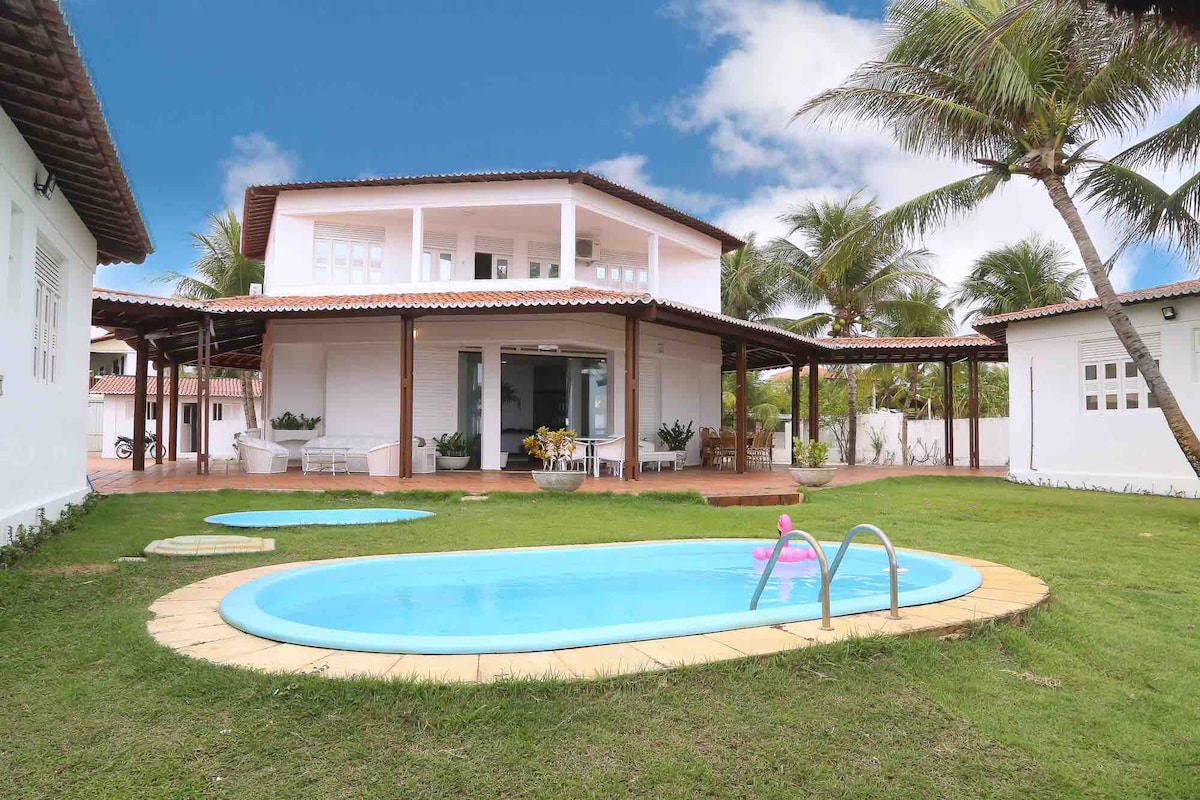 The exterior of a two-story house is showcased, surrounded by a well-maintained lawn. A small pool with steps can be seen in the foreground, alongside comfortable seating areas under a spacious covered patio. Lush palm trees add to the tropical atmosphere.