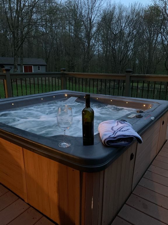 A six-person jacuzzi is situated on a wooden deck, surrounded by trees. Soft lighting highlights the bubbling water. A wine bottle rests on the edge, accompanied by an empty glass and a folded towel, creating a tranquil atmosphere for relaxation.