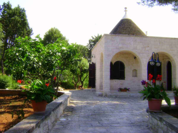 A charming stone structure features a conical roof and arched entrances. Lush greenery and colorful flowers in decorative pots adorn the pathway, leading to the entrance. Trees and shrubs create a peaceful outdoor setting, enhancing the tranquil atmosphere of the surroundings.