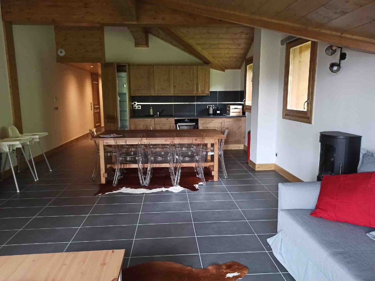 A spacious living area features wooden beams and tile flooring. A large dining table with clear chairs is positioned near the kitchen, which is partially visible in the background. A cozy seating area includes a light grey sofa with a red accent pillow, enhancing the overall design.