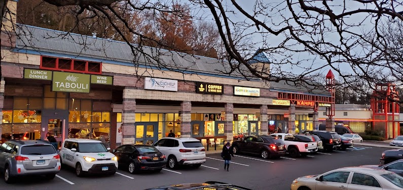 A shopping center exterior is visible in the evening light, showcasing multiple storefronts with illuminated signs. A variety of vehicles occupy the parking lot, while a few individuals walk toward the entrance. The overall environment appears welcoming and active.