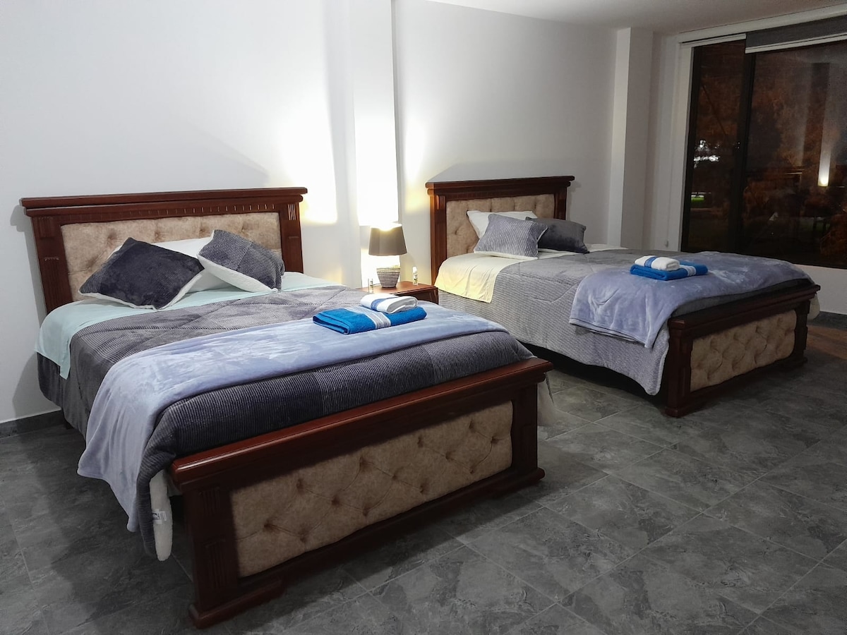 The room features two spacious beds, each adorned with neatly arranged pillows and soft blankets. A nightstand with a lamp provides lighting, while a large window allows natural light to fill the space, complemented by modern gray tiled flooring.