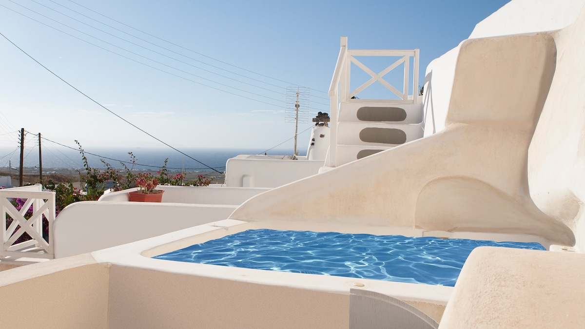 An outdoor jacuzzi is featured on a terrace, surrounded by smooth, white walls and scenic views of the sea. Steps lead upward, providing access to additional outdoor areas. Soft blue water contrasts with the neutral tones of the surrounding architecture.