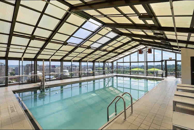 An indoor pool area is featured with large glass panels allowing ample natural light. The clear water reflects the contemporary architecture, and lounge chairs line one side. Scenic views of the surrounding cityscape are visible through the expansive windows, offering a bright and airy atmosphere.