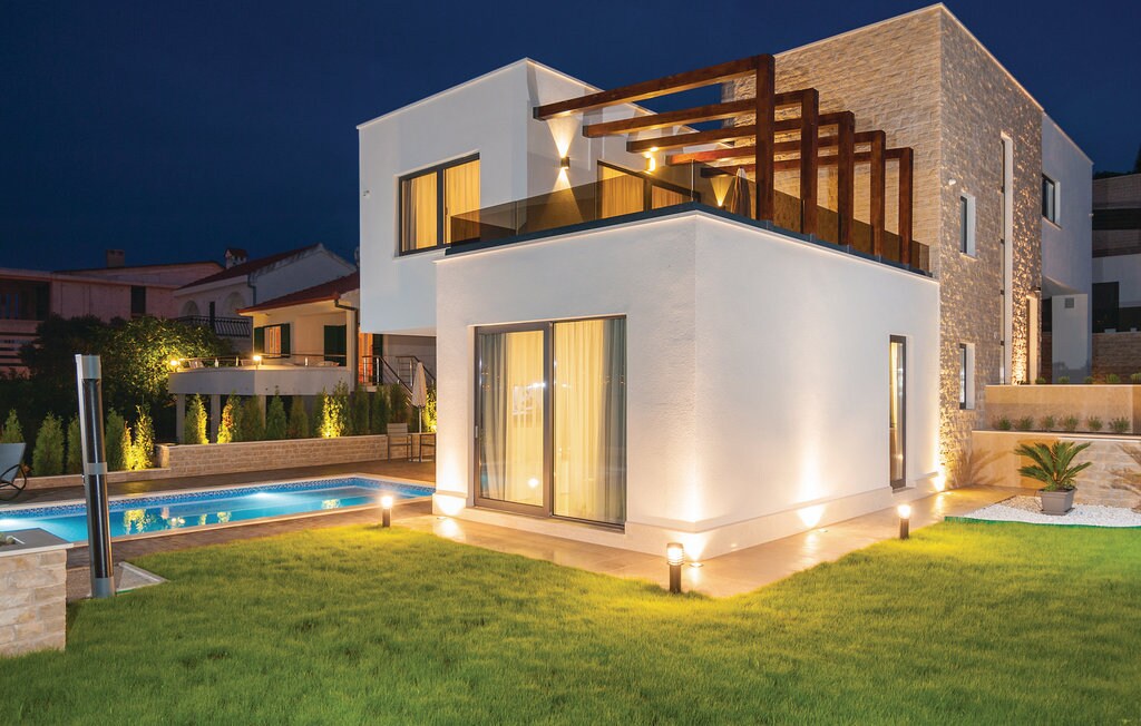 The villa's modern exterior is illuminated at night, showcasing a unique architectural design with large glass windows and a spacious terrace. Surrounding areas feature manicured lawns and a swimming pool, enhancing the overall aesthetic of the property while inviting outdoor relaxation.