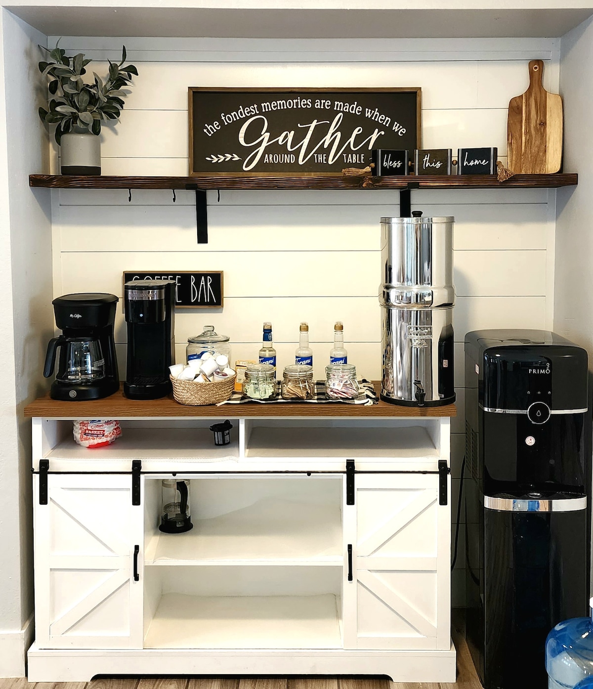 A dedicated coffee bar area features a coffee maker and an electric kettle on a sturdy wooden countertop. Shelves display decorative items and a sign that emphasizes gathering. A variety of bottled water and drink options are arranged in a woven basket, all presented against a light-colored wall.