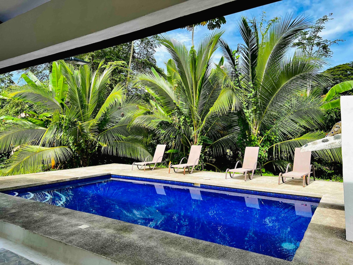 A private pool is positioned in the foreground, surrounded by a sun terrace featuring four loungers. Lush green palm trees create a tropical backdrop, while clear skies and gentle lighting enhance the serene outdoor setting.