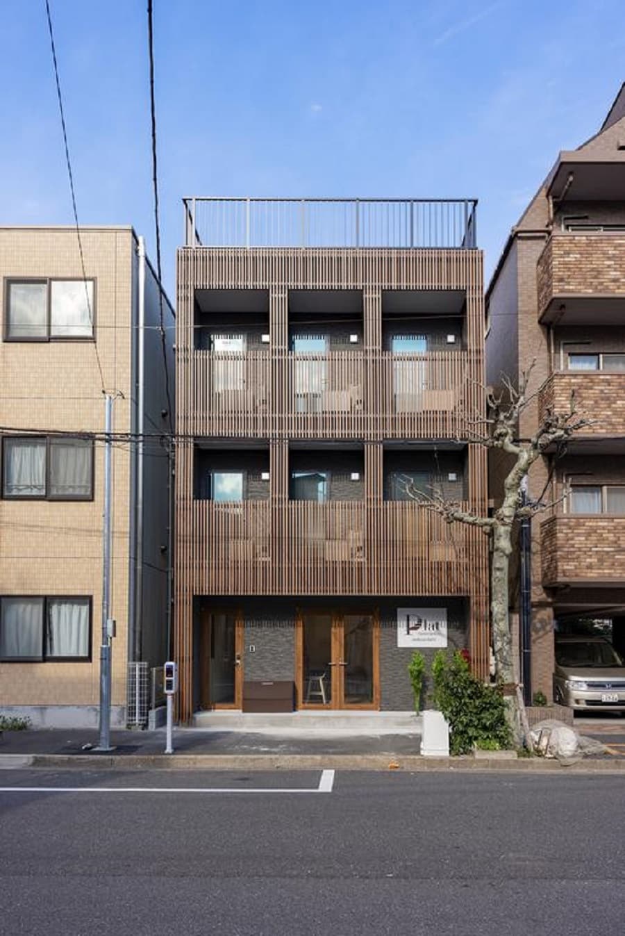 The exterior of a modern four-story building is shown, featuring a wooden facade with slatted screens. Ground-level entrance is visible, along with large windows on each floor providing light. Surrounding structures are present, contributing to the urban setting.