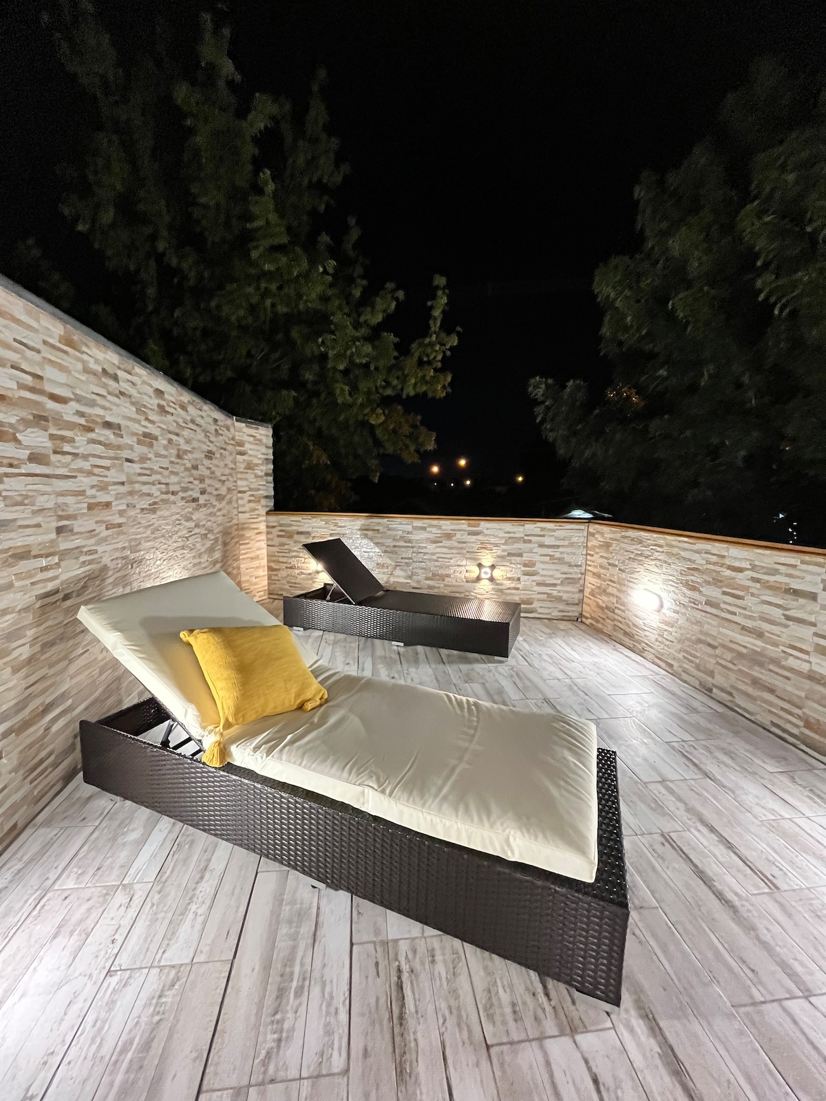 Outdoor terrace features two sun loungers arranged alongside a stone wall, providing a quiet space for relaxation. Soft lighting enhances the seating area, and natural greenery is visible in the background, creating a serene environment.