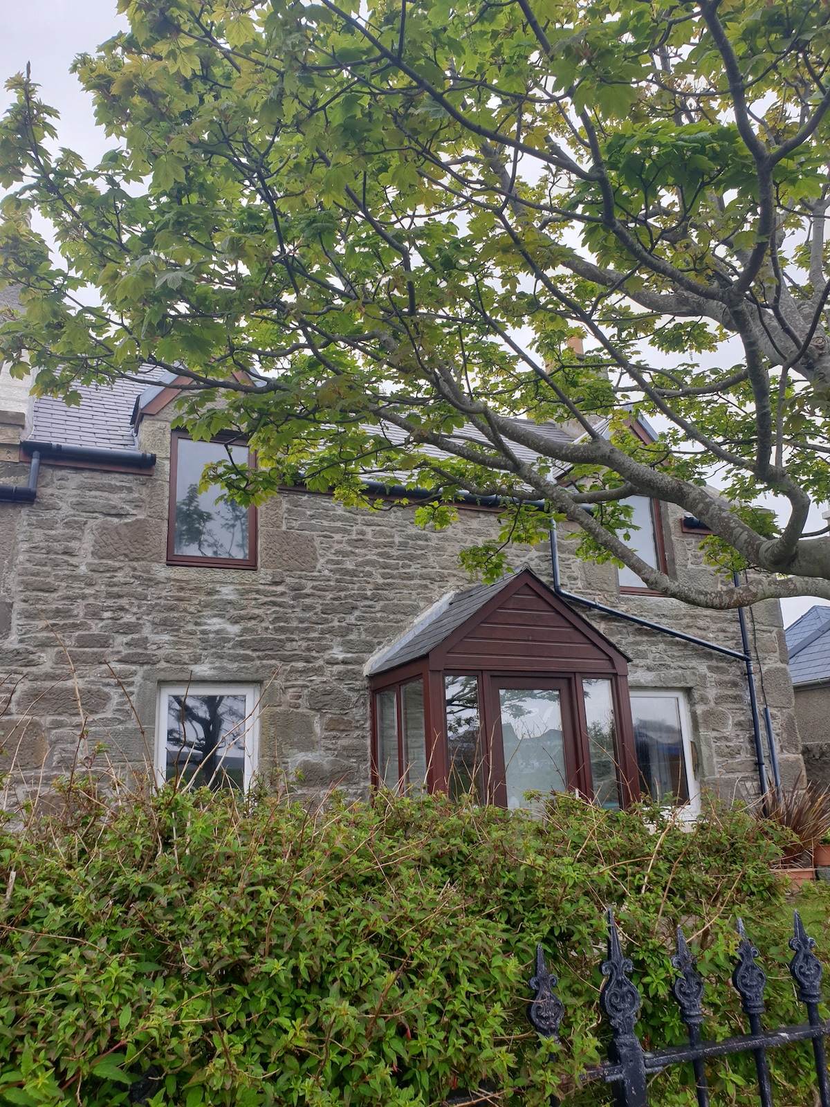 An exterior view of the stone-built apartment features large windows surrounded by a lush green garden. A decorative tree provides shade, while the building maintains a blend of traditional and modern architectural elements.