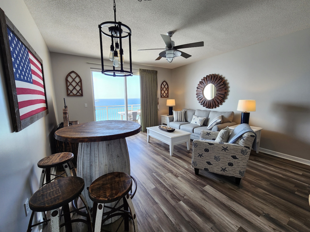 A bright and spacious living area features a circular wooden table with three stools, alongside a comfortable couch and decorative pillows. A large mirror reflects light, while a wall showcases a framed American flag. Windows provide views of the ocean beyond.