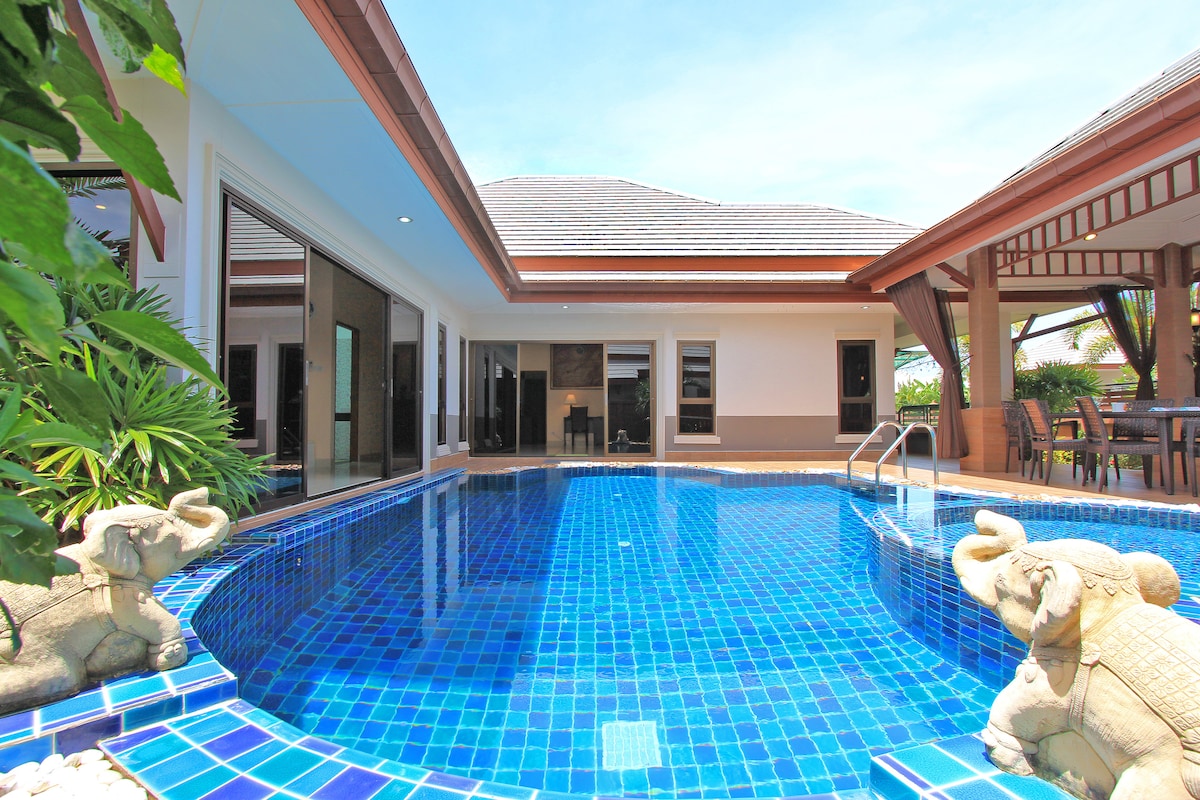 A private pool area is surrounded by lush greenery, featuring a tiled pool with a vibrant blue hue. Two decorative elephant statues are positioned at the edges of the pool. Spacious lounge areas are visible beyond the water, inviting relaxation and enjoyment.