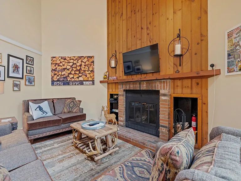 A cozy living area features rich wooden paneling and a large stone fireplace. Comfortable seating is arranged around a rustic coffee table, enhancing the warm atmosphere. A flat-screen television is mounted above the fireplace, with framed art and a stack of firewood displayed nearby.
