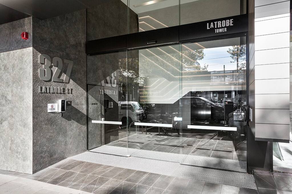The entrance to Latrobe Tower is depicted, featuring large glass doors that provide a clear view of the interior lobby. A sleek design with modern finishes is evident, and the building name is prominently displayed. Natural light brightens the entrance area.