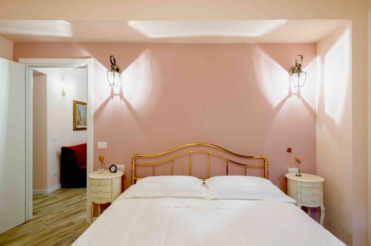The bedroom features a spacious bed with a delicate gold frame against soft pink walls. Two nightstands with lamps are placed on either side of the bed, while light is softly reflected, creating a warm ambiance.