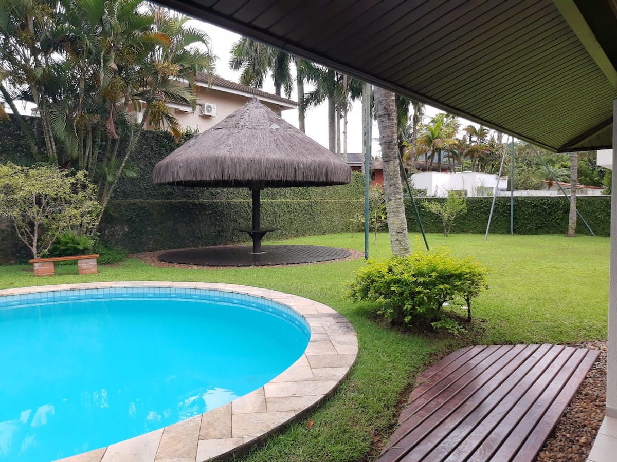An inviting outdoor space is highlighted by a sparkling blue pool and a thatched-roof kiosk set on a wooden platform. Lush green grass surrounds the area, featuring a swinging bench and carefully maintained landscaping that enhances the tranquil environment.