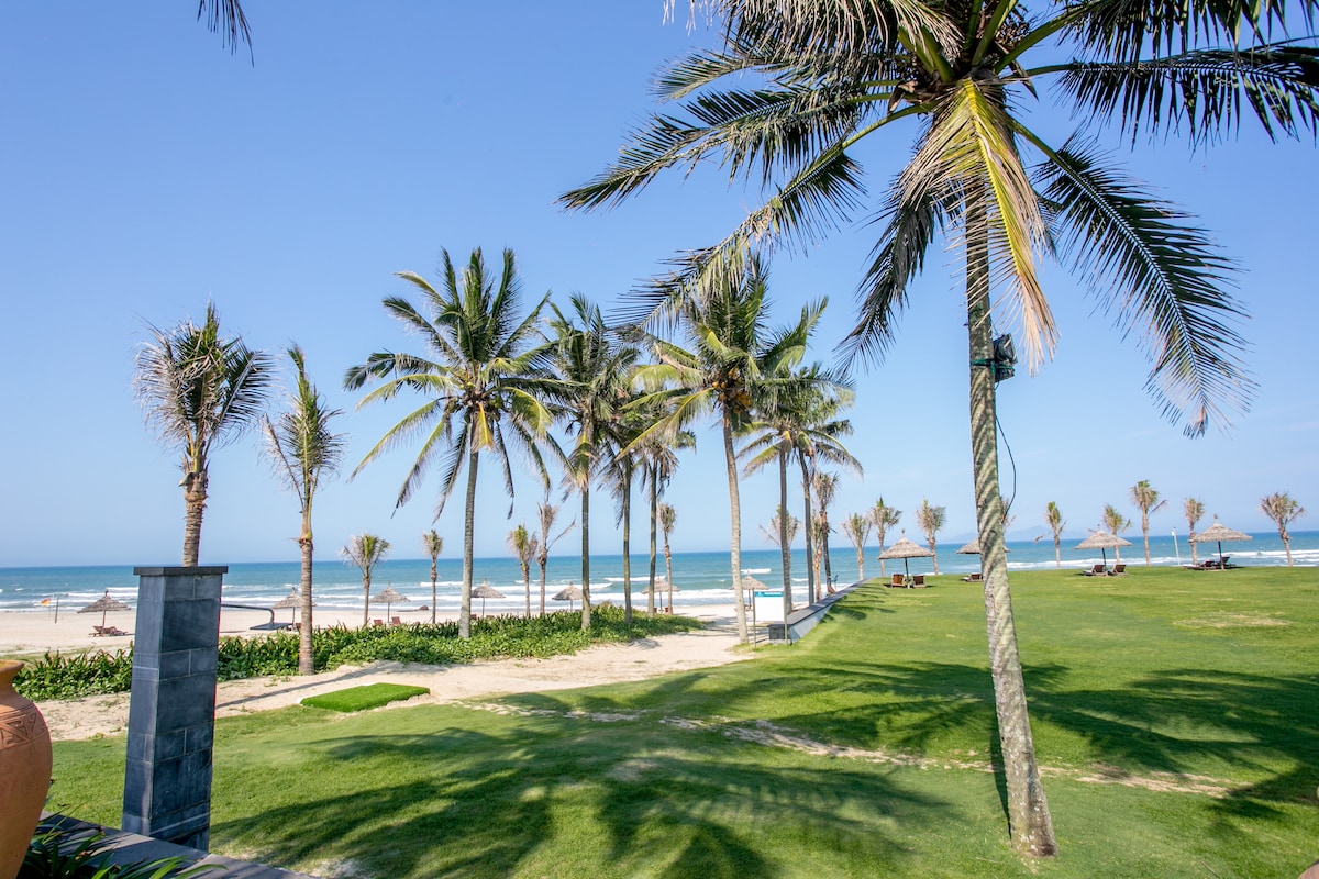 Lush green lawns are bordered by swaying palm trees, leading to a sandy beach. Clear blue skies stretch above, with the ocean waves gently lapping at the shore. Sun loungers are positioned in the background, providing a relaxing spot to enjoy the beach view.