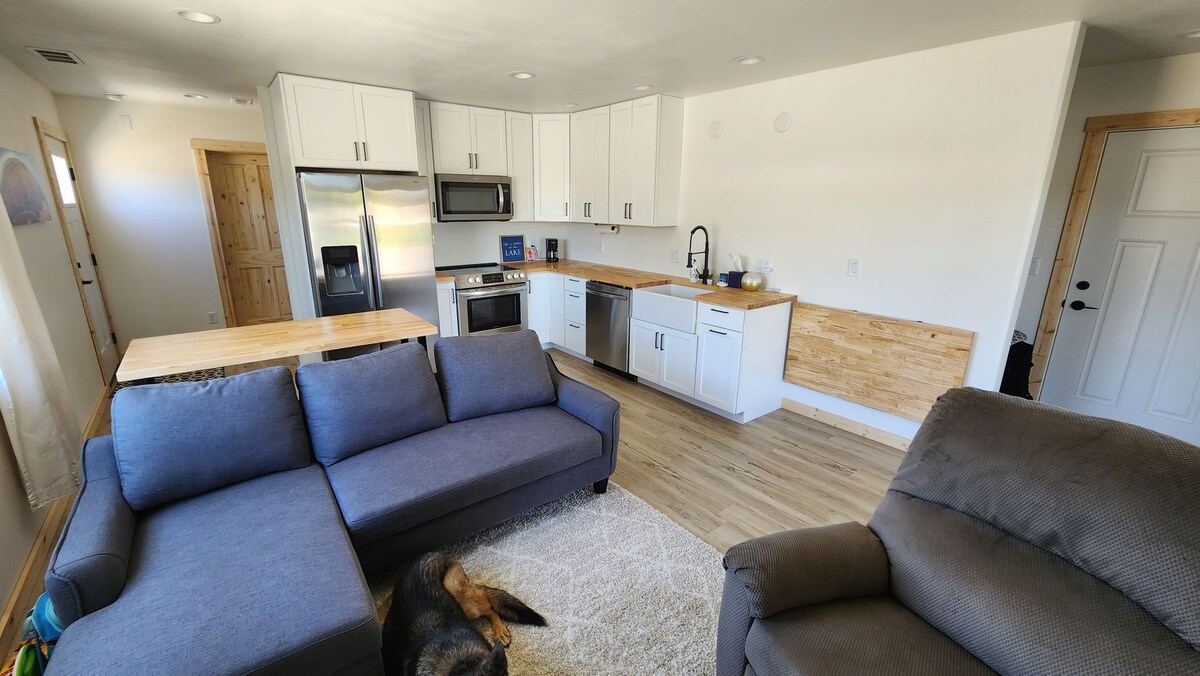 A modern kitchen area is shown, featuring stainless steel appliances and a light wood countertop. A spacious sofa with a cozy texture is positioned nearby, alongside a comfortable recliner. Natural light fills the room, enhancing the clean lines of the cabinetry and open floor plan.