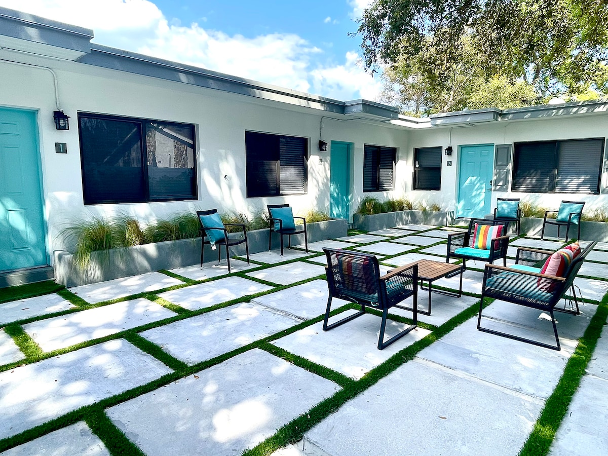 A private courtyard features a clean layout with square concrete tiles and green grass patches. Several chairs are arranged around a central wooden table, creating a gathering space. Bright blue doors and large windows enhance the inviting appearance of the space.