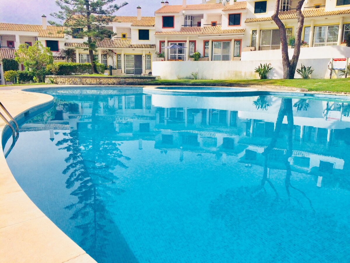 A sparkling pool reflects the surrounding residential buildings, creating a serene atmosphere. Light blue tiles line the pool's edge, and lush greenery is visible in the background. The area is framed by well-kept landscaping, providing a calm outdoor setting.