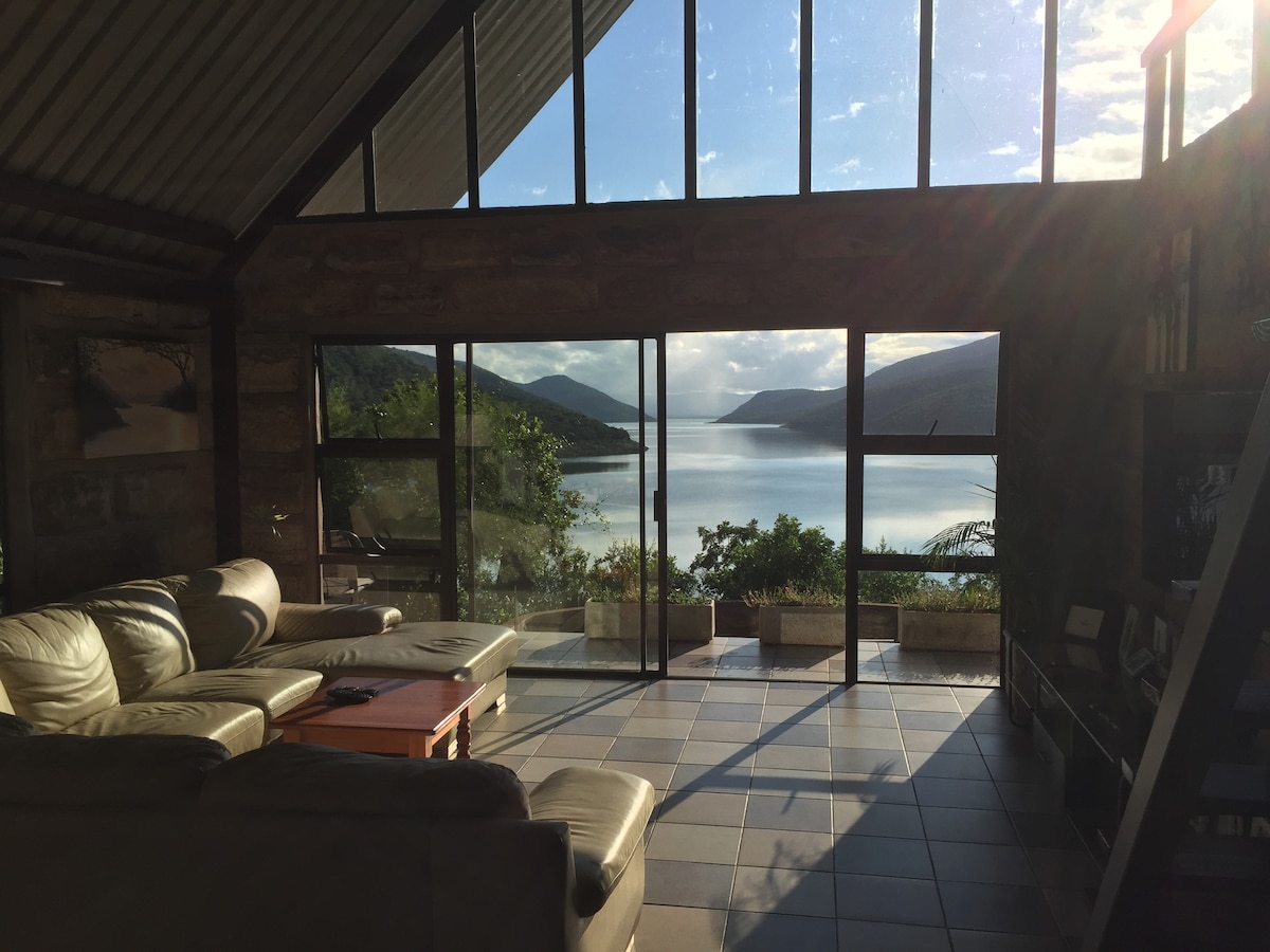 An open living area features large glass walls, providing stunning views of Jozini Dam and the surrounding landscape. Comfortable seating arrangements include a light-colored sofa, and natural light fills the space, creating a bright and airy atmosphere.